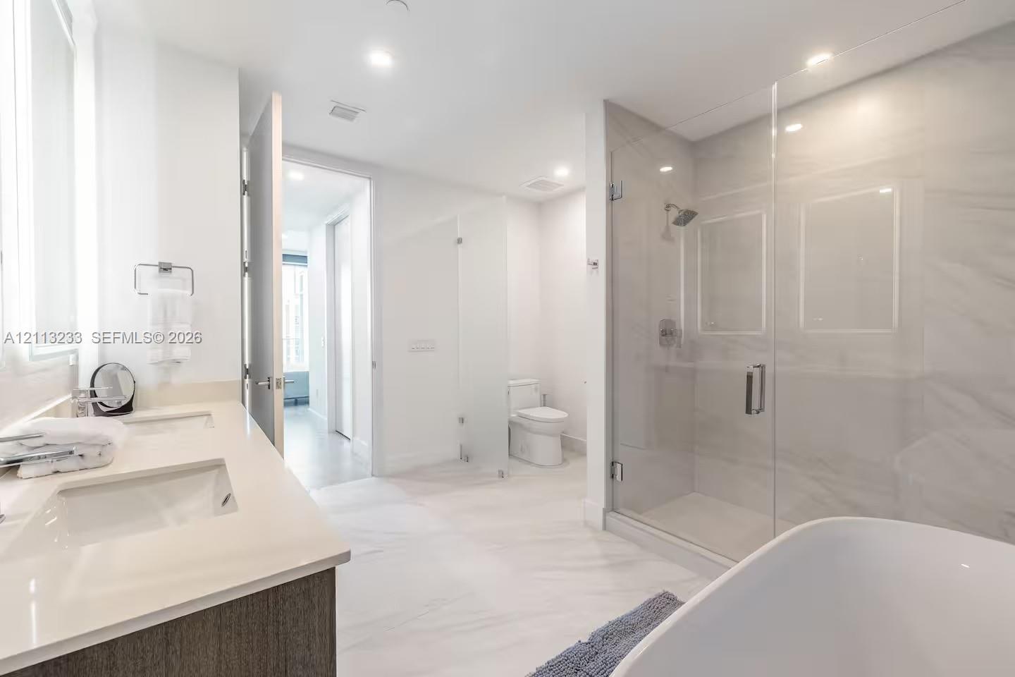 Dual-sink vanity with light-toned countertop, freestanding soaking tub, glass-enclosed walk-in shower with tiled surround, and separate enclosed water