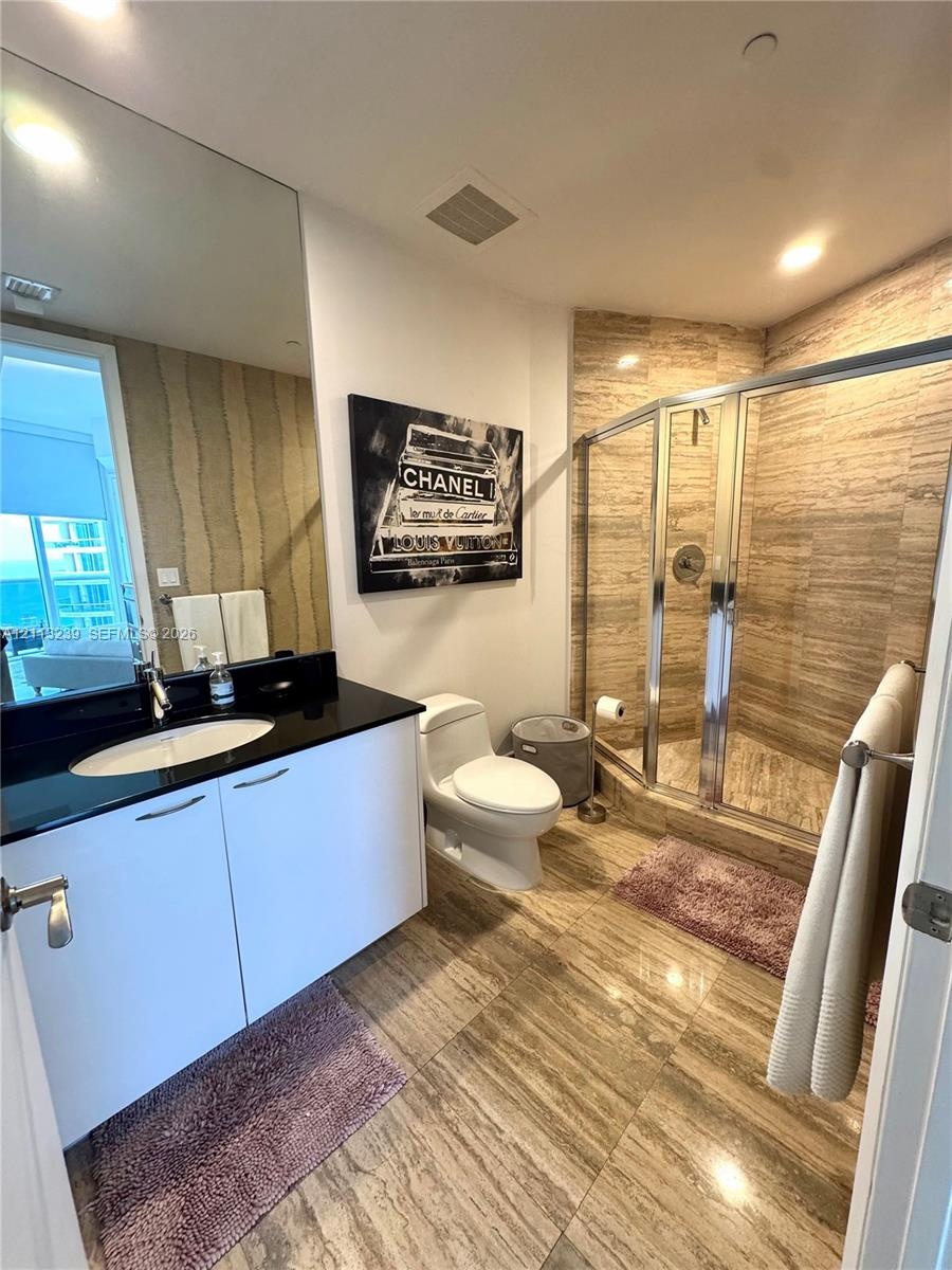Full-height vanity mirror, dark stone-surface countertop, white cabinetry, walk-in shower with stone tile surround, and large-format stone floor tile