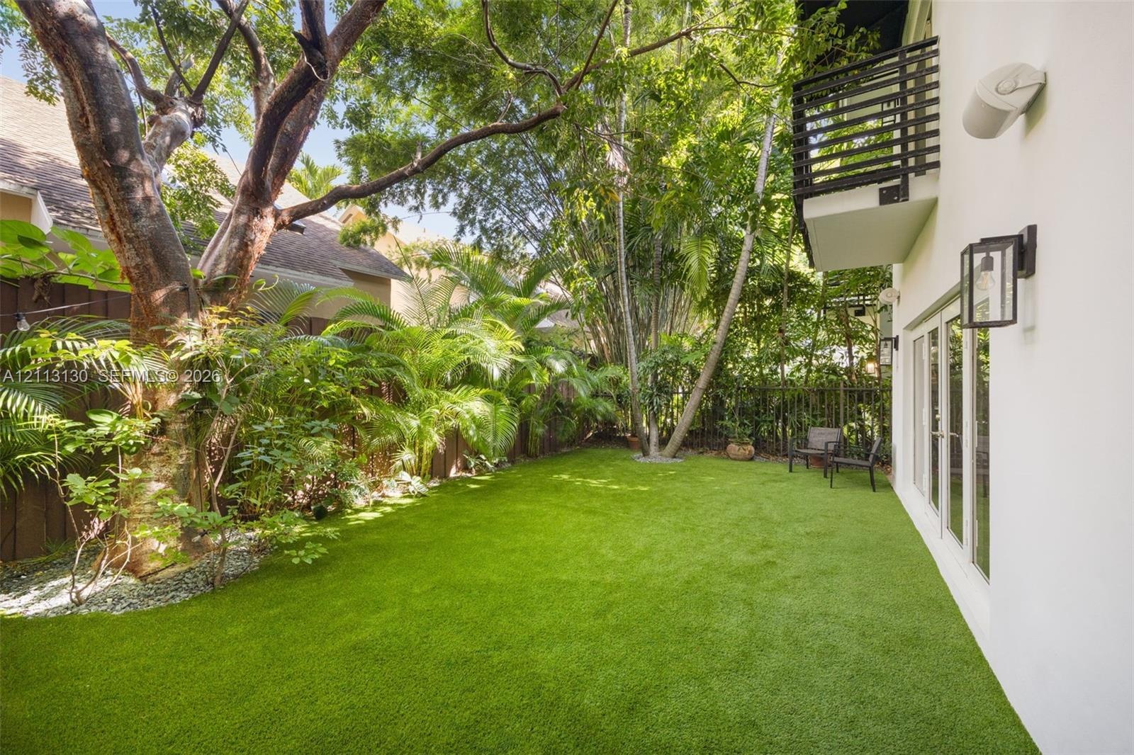Private backyard featuring synthetic turf, mature trees, wood privacy fencing, a second-story balcony with metal railings, and exterior wall-mounted l