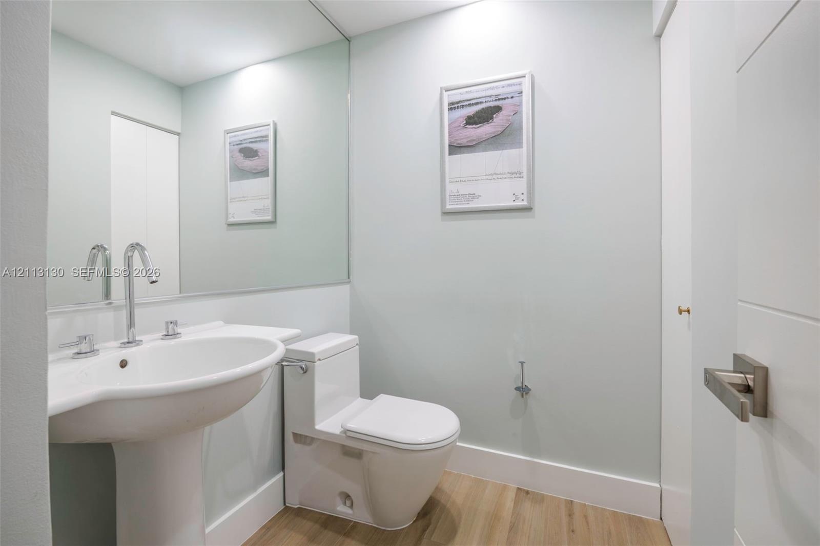 Pedestal sink with high-arc polished chrome faucet, expansive wall-mounted mirror, one-piece dual-flush toilet, and wood-finish flooring