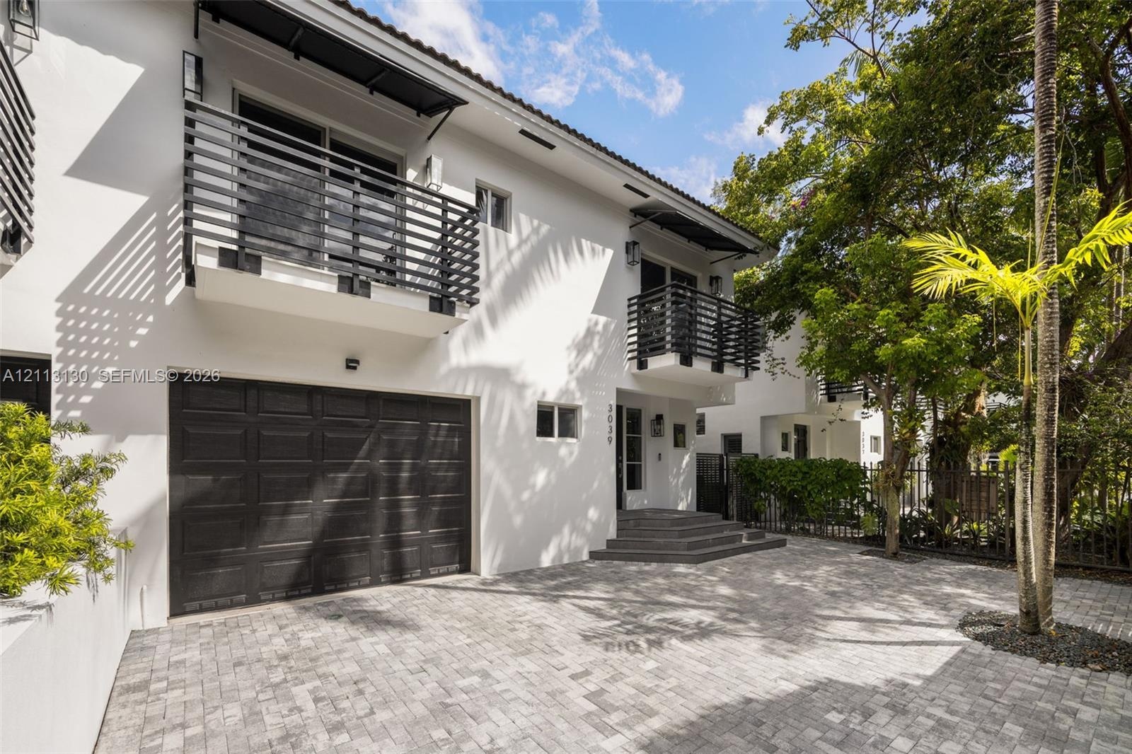 Exterior featuring smooth white stucco siding, dual second-story balconies with horizontal slat railings, a paved stone driveway, and an attached mult