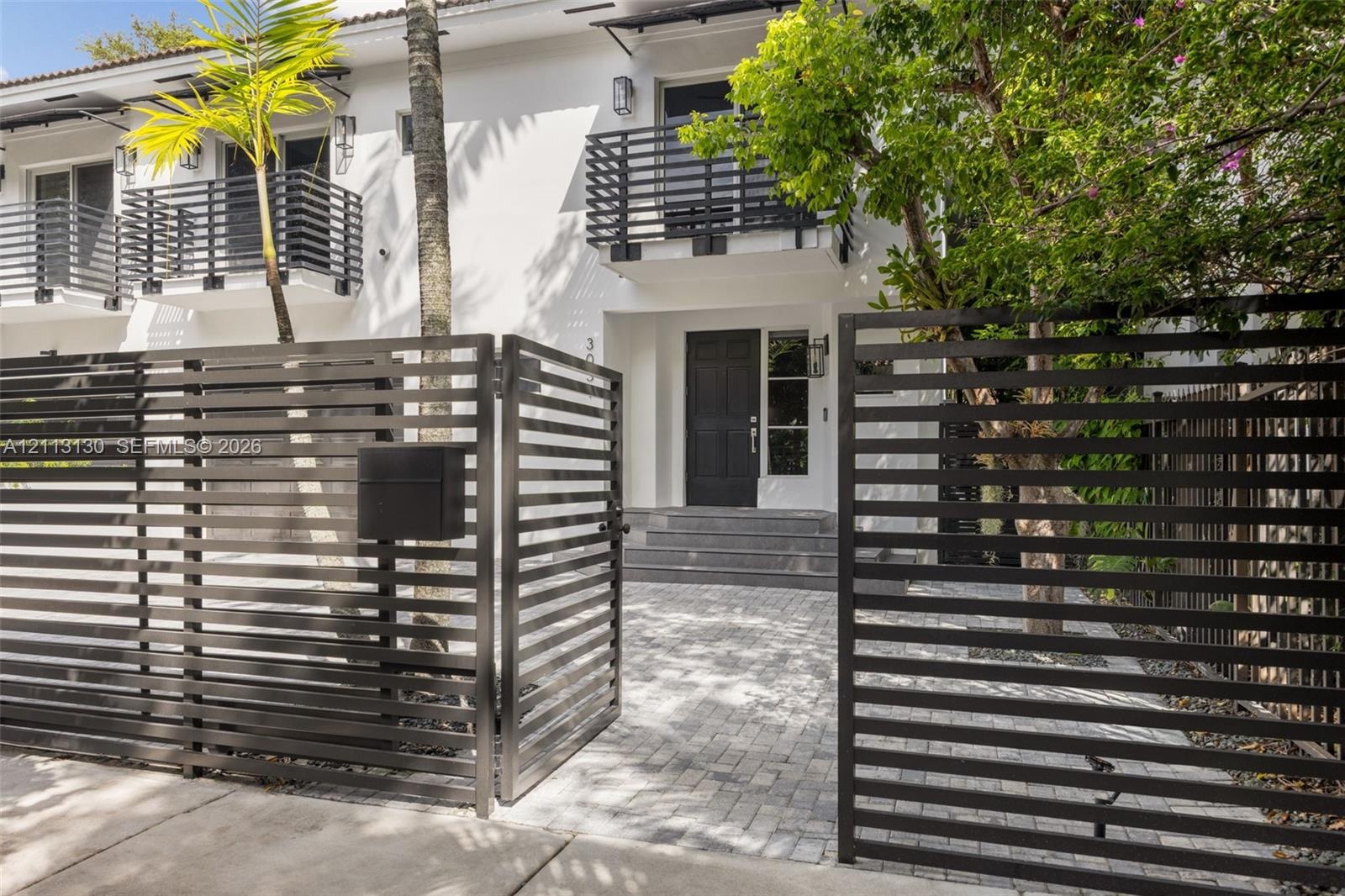 Exterior facade featuring a paved stone driveway, horizontal slat metal privacy fencing, solid panel entry door, and upper-level balconies with matchi