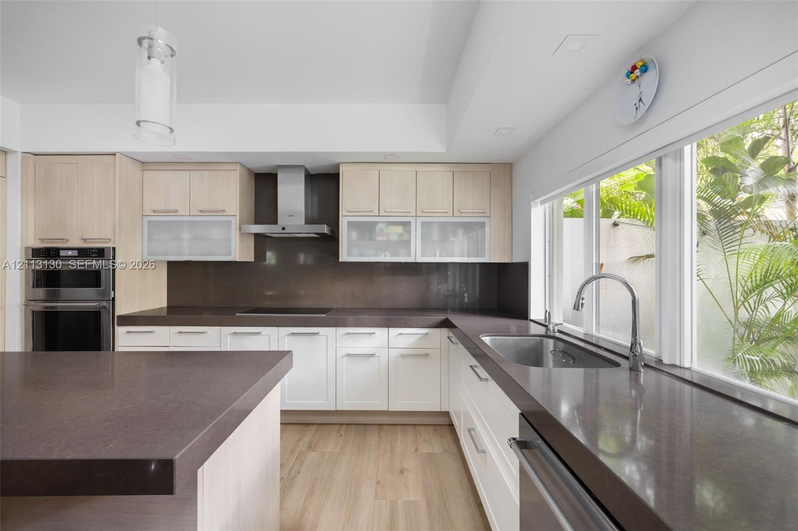 Kitchen featuring light wood-finish cabinetry, solid-surface countertops, a stainless steel chimney-style range hood, built-in double ovens, and a dua