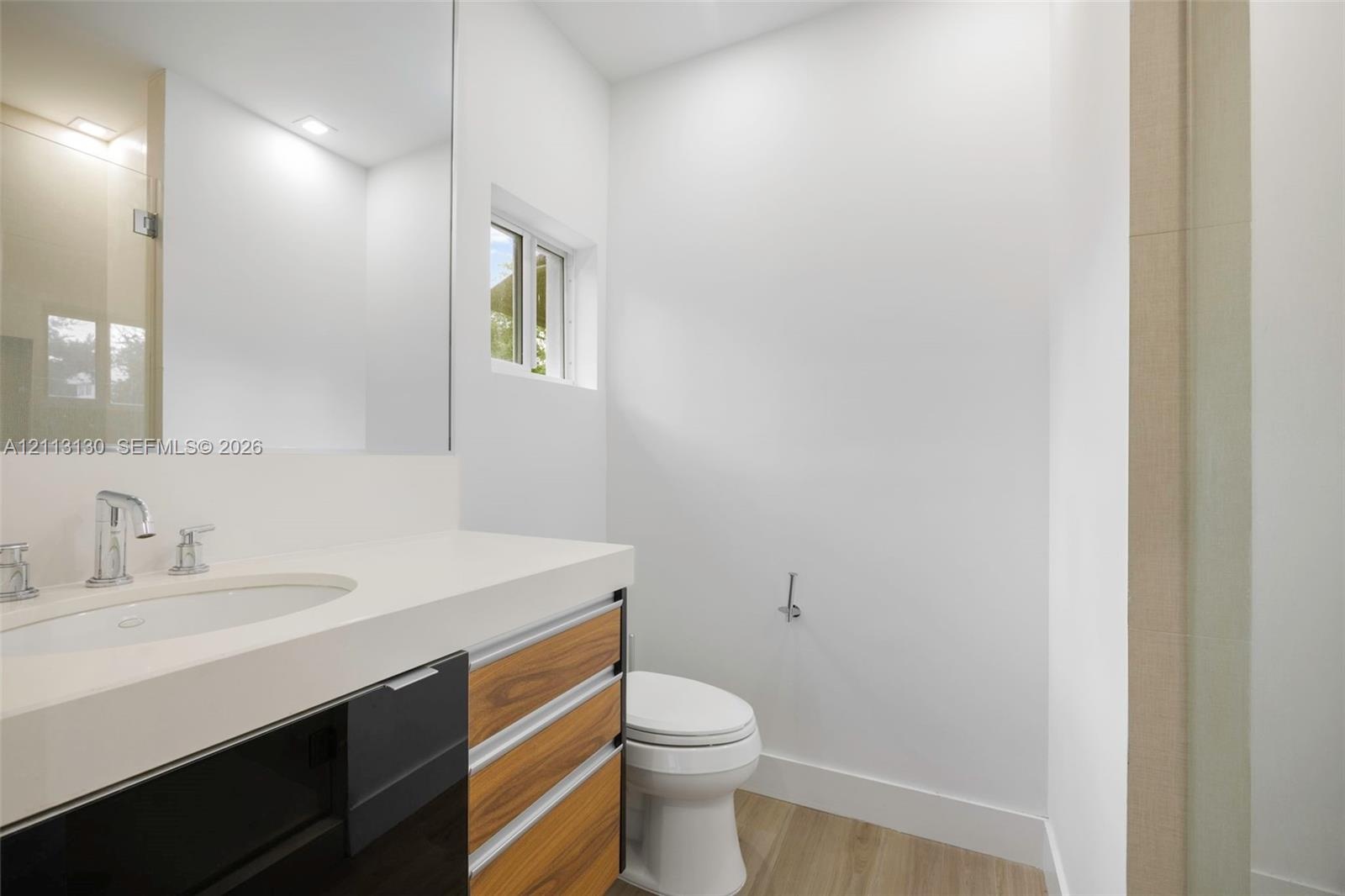 Wall-mounted mirror, vanity with wood-finish cabinetry and light-toned countertop, integrated basin, chrome-finish faucet, and wood-finish flooring