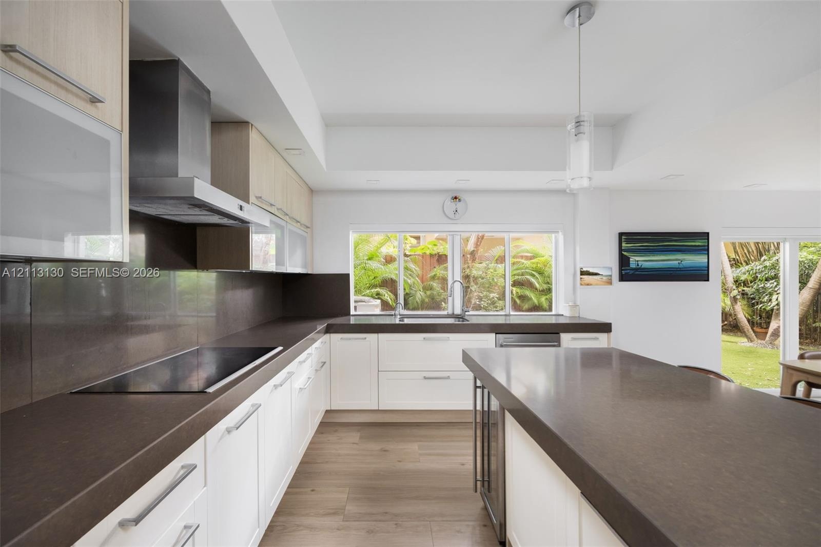 Modern kitchen featuring white flat-panel cabinetry, dark stone-finish countertops, stainless steel range hood, built-in cooktop, and wood-finish floo