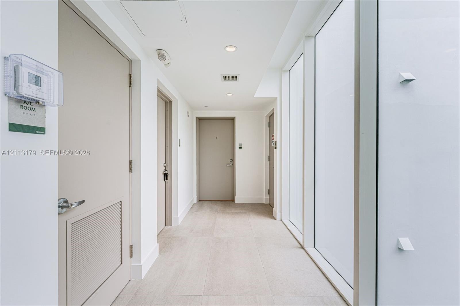 Interior corridor featuring large-format tile flooring, floor-to-ceiling glass wall panels, recessed lighting, and solid-core doors
