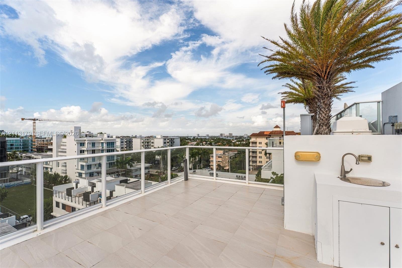 Rooftop terrace featuring light-toned tile flooring, a white glass-paneled railing, integrated wet bar with a stainless steel sink, and mature palm tr