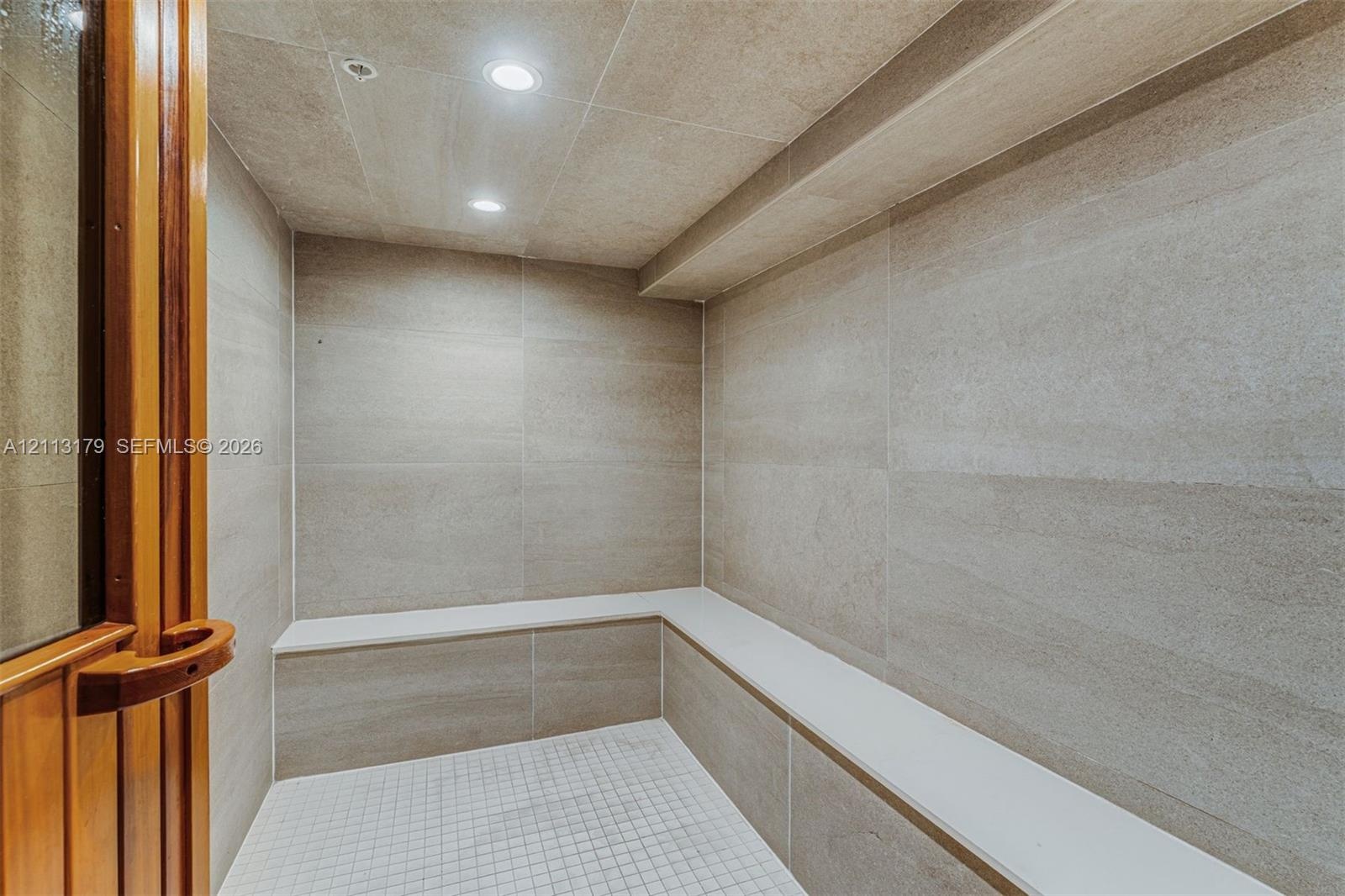 Steam room featuring large-format stone-tile walls, built-in tiered stone seating, mosaic-tile flooring, and recessed lighting behind a wood-framed gl