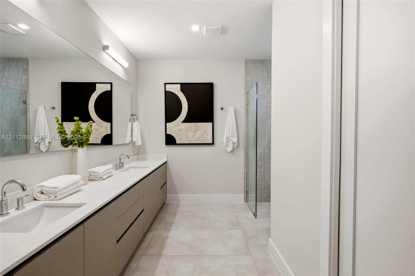 Dual-sink floating vanity with neutral cabinetry, expansive wall-mounted mirror, light-toned stone-finish flooring, and glass-enclosed shower with ful