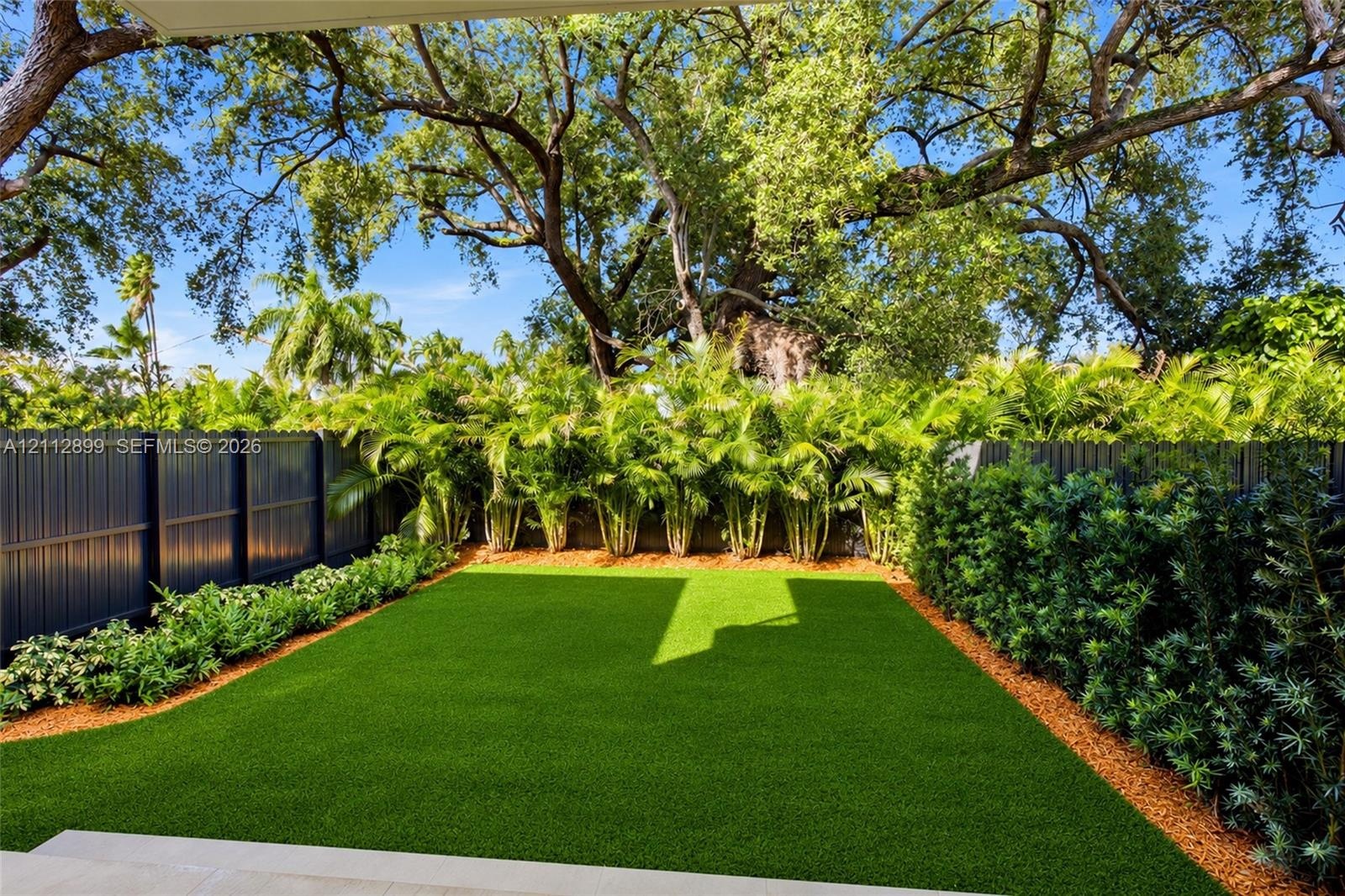 Backyard featuring artificial turf, perimeter fencing, mature canopy trees, and dense foliage border