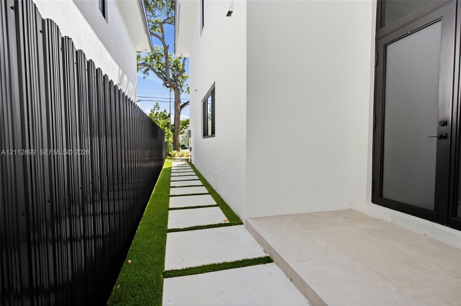 Side exterior featuring white stucco siding, a concrete paver walkway set in artificial turf, a black metal corrugated privacy fence, and a glass-pane