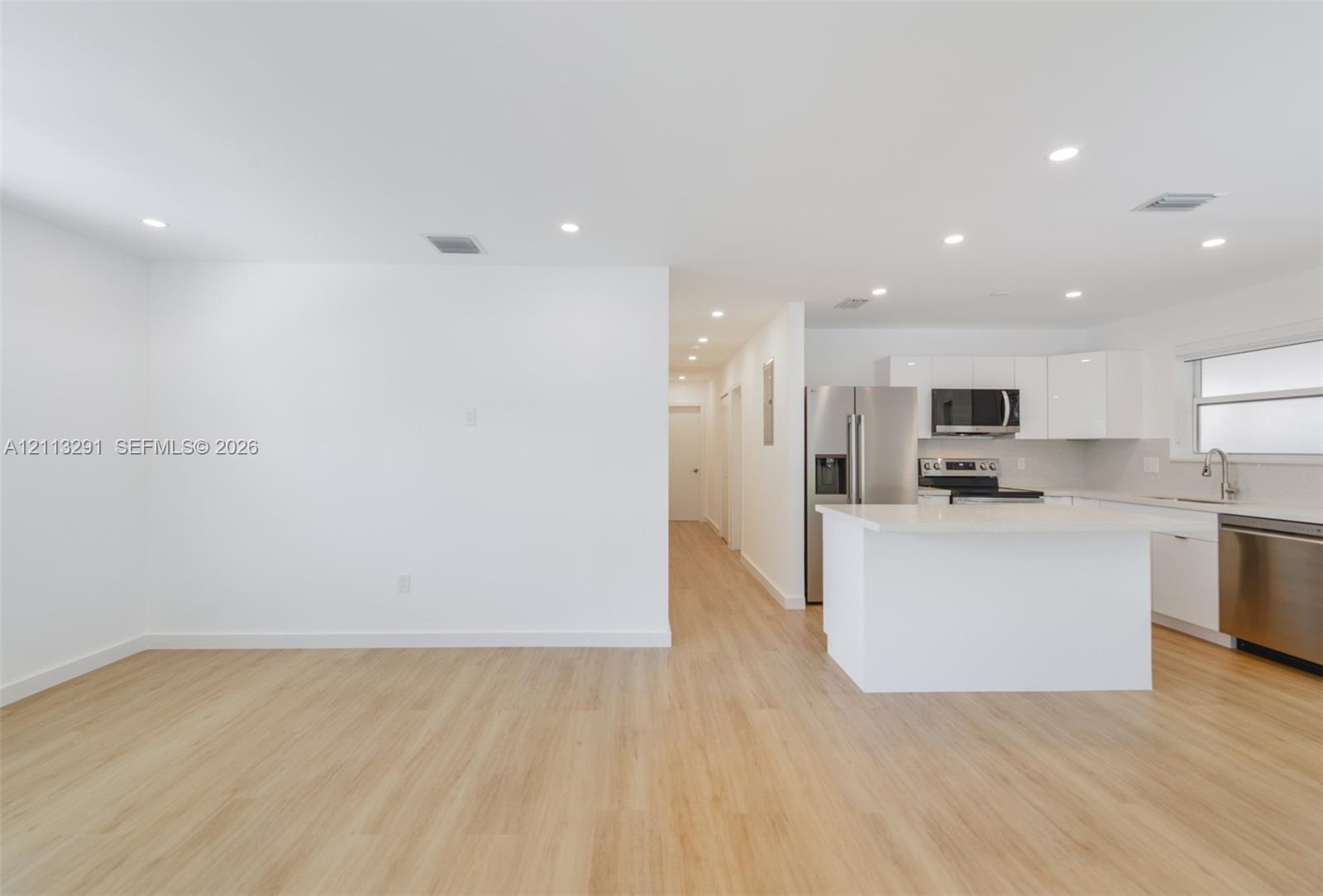 Open-concept interior featuring light wood-finish flooring, recessed lighting, white gloss cabinetry, quartz countertops, and stainless steel applianc