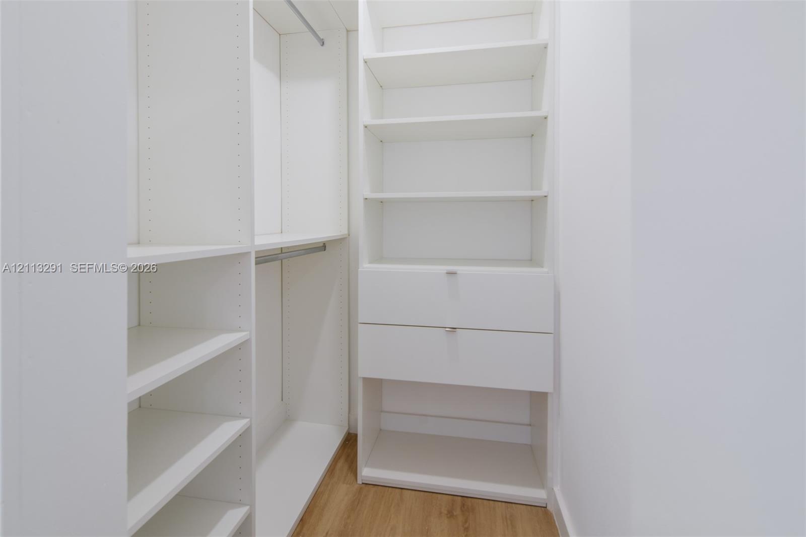 Built-in white storage system featuring adjustable shelving, integrated hanging rods, and dual drawers with silver-tone hardware over wood-finish floo