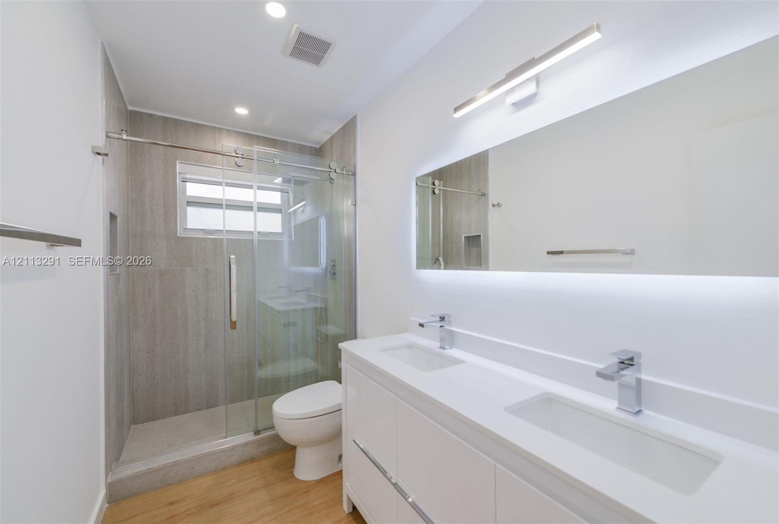 Double vanity with a white countertop and rectangular undermount sinks, illuminated mirror with integrated backlighting, wood-finish flooring, and a w