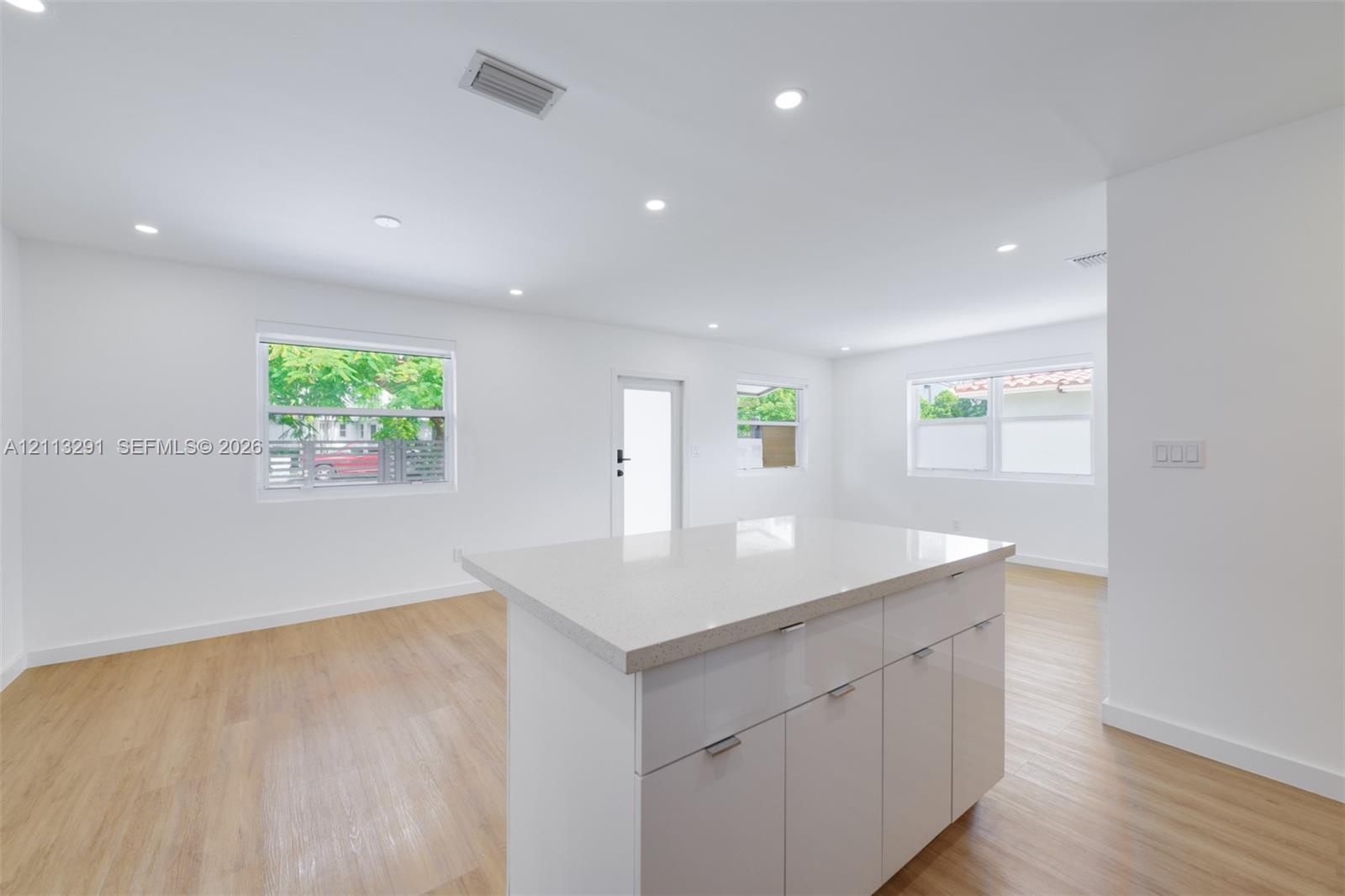 Open-concept interior featuring wood-finish flooring, recessed lighting, and a central island with a stone-composite countertop and high-gloss cabinet