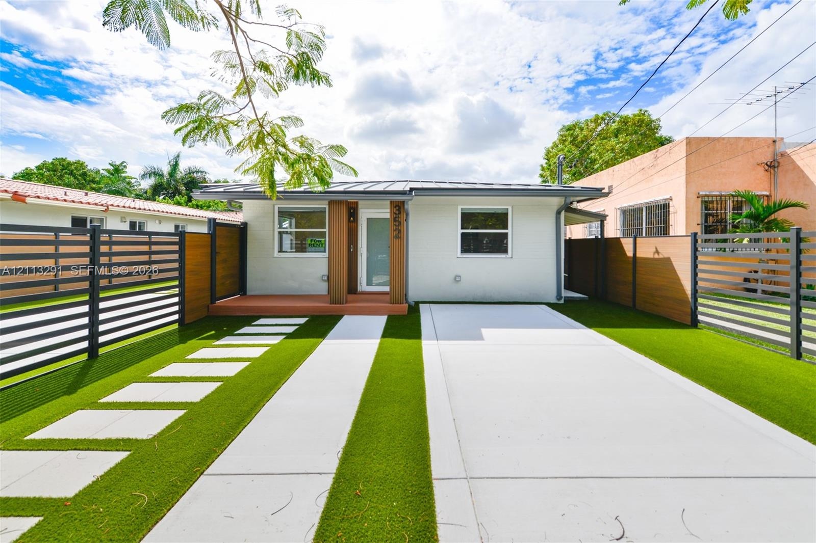 Single-story exterior featuring a metal roof, concrete driveway, synthetic turf lawn, stone paver walkway, and horizontal slat privacy fencing