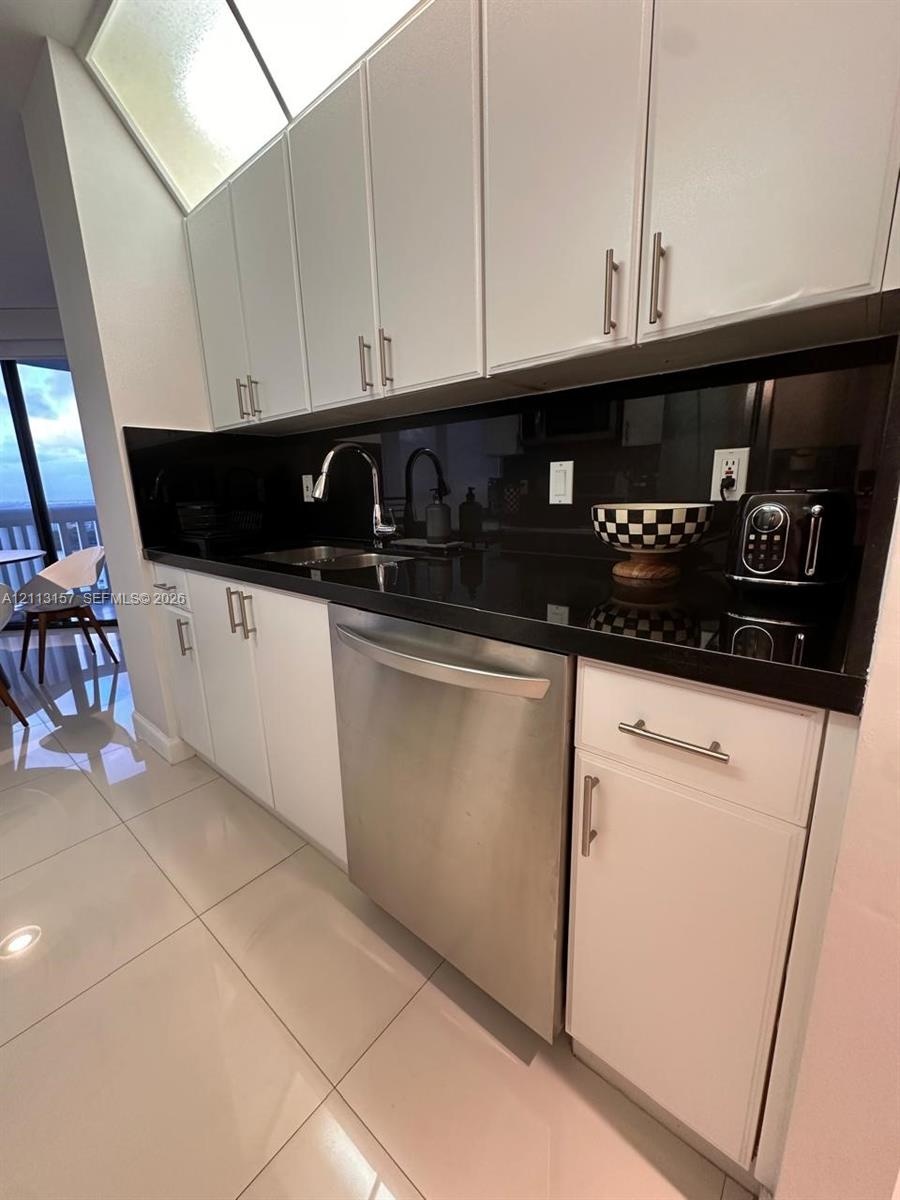 Kitchen featuring white cabinetry with metallic pulls, dark solid-surface countertops, a stainless steel dishwasher, a sleek gooseneck faucet, and lig