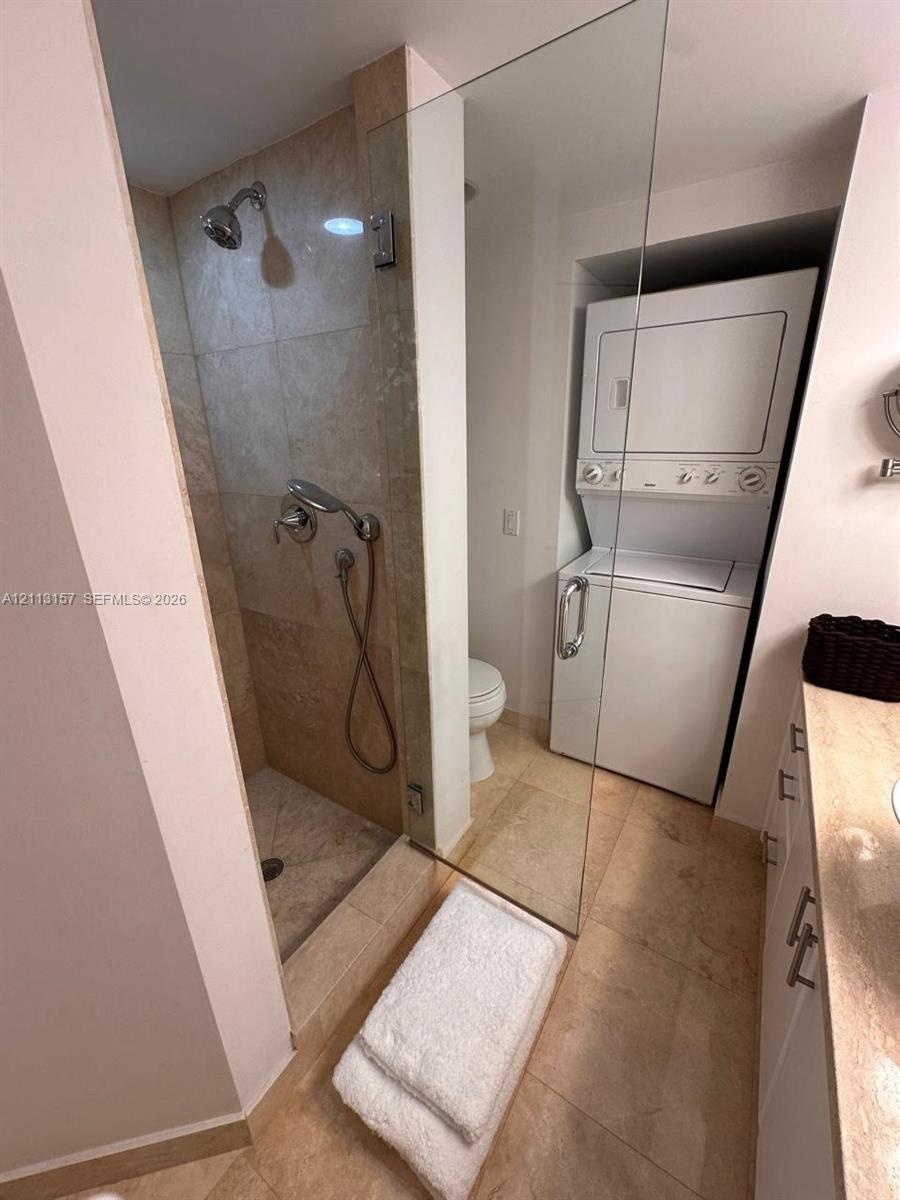 Walk-in shower with stone-finish tile surround and glass enclosure, integrated laundry unit with stacked washer and dryer, and vanity with stone-finis