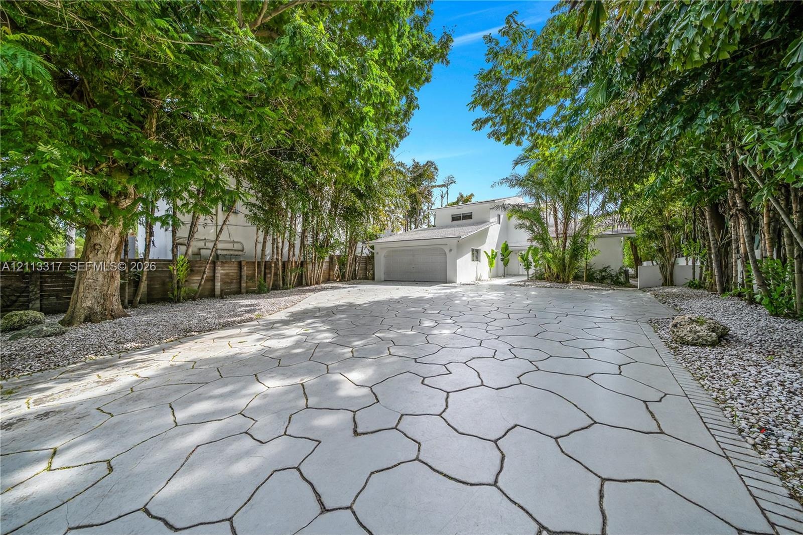 Extensive paver driveway featuring a decorative flagstone pattern, mature perimeter trees, stone-accented landscaping, and an attached garage
