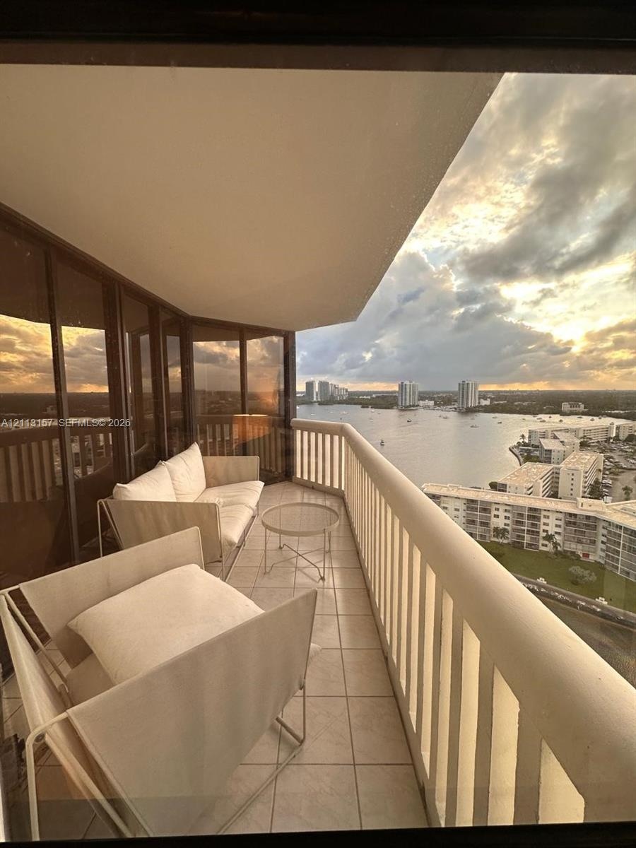 Covered balcony featuring expansive glass paneling, tile flooring, and a white vertical railing, overlooking an expansive water landscape and urban sk