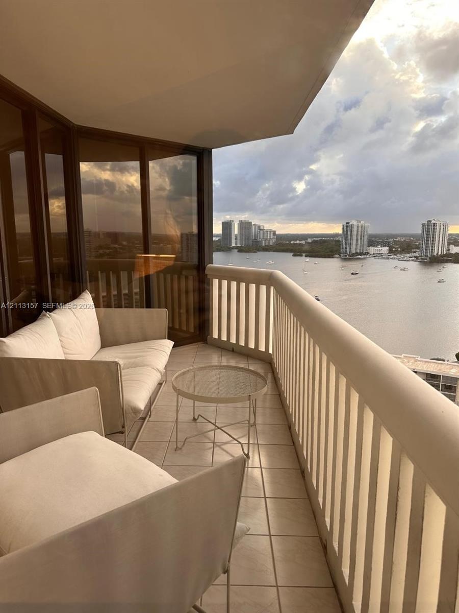 Covered balcony featuring tile flooring, metal railings, and floor-to-ceiling tinted glass panels overlooking the water and urban skyline