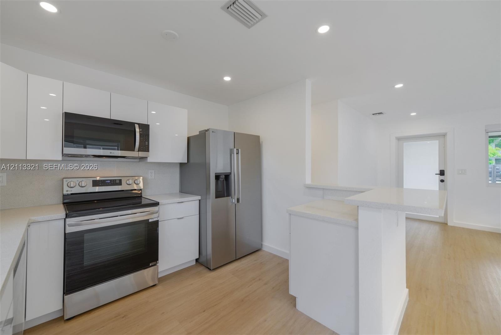 Modern kitchen featuring white flat-panel cabinetry, light-toned countertops, stainless steel appliances, wood-finish flooring, and recessed lighting