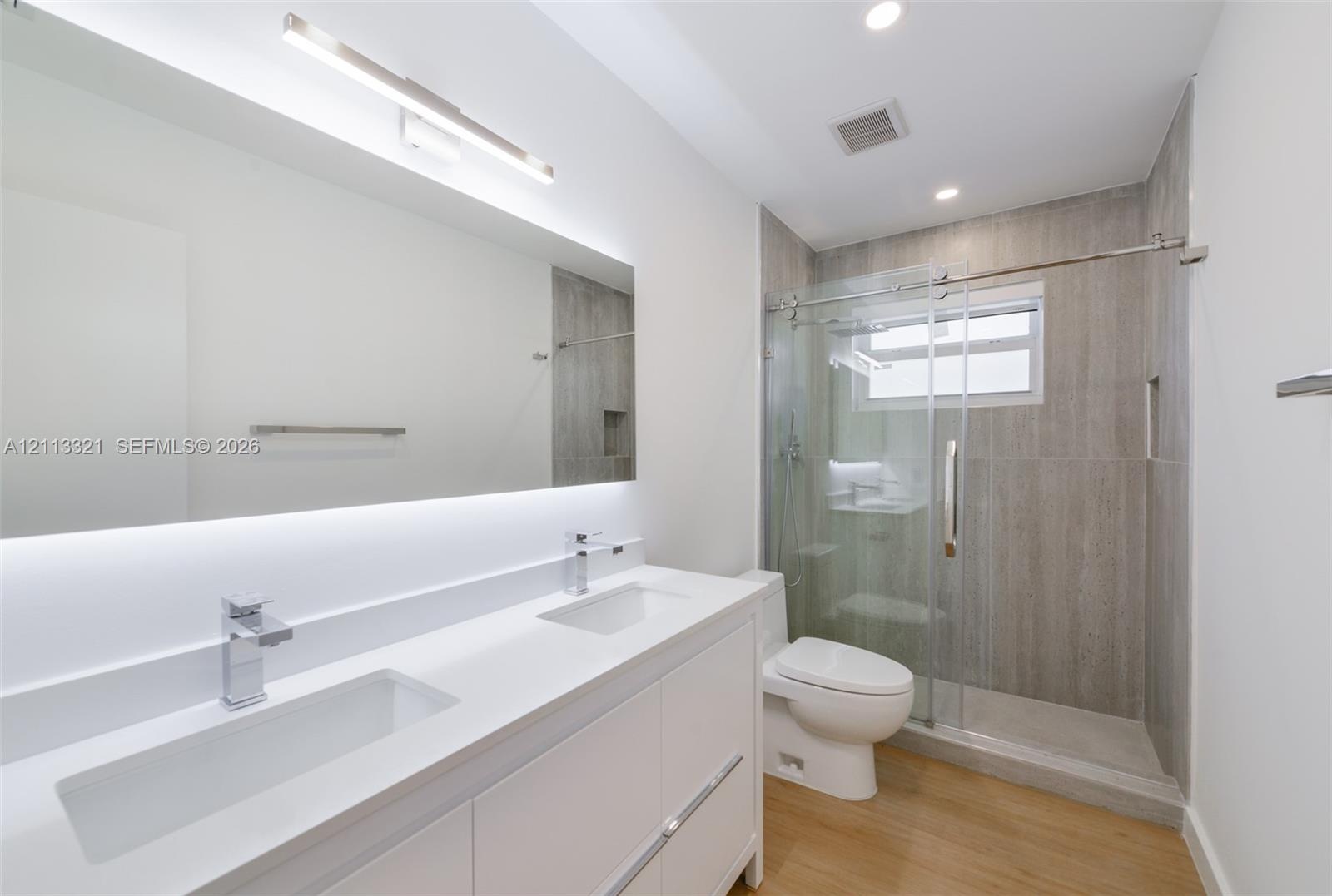 Dual-sink vanity with white stone-finish countertop, under-mount sinks, chrome faucets, full-width mirror with integrated lighting, and glass-enclosed