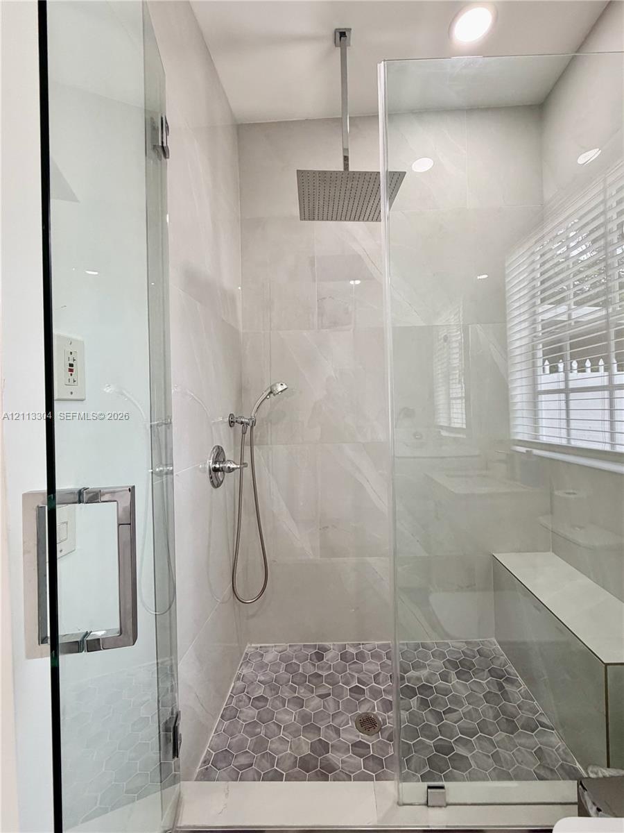 Walk-in shower featuring a frameless glass enclosure, ceiling-mounted rainfall showerhead, handheld sprayer, built-in bench, and grey hexagonal floor 