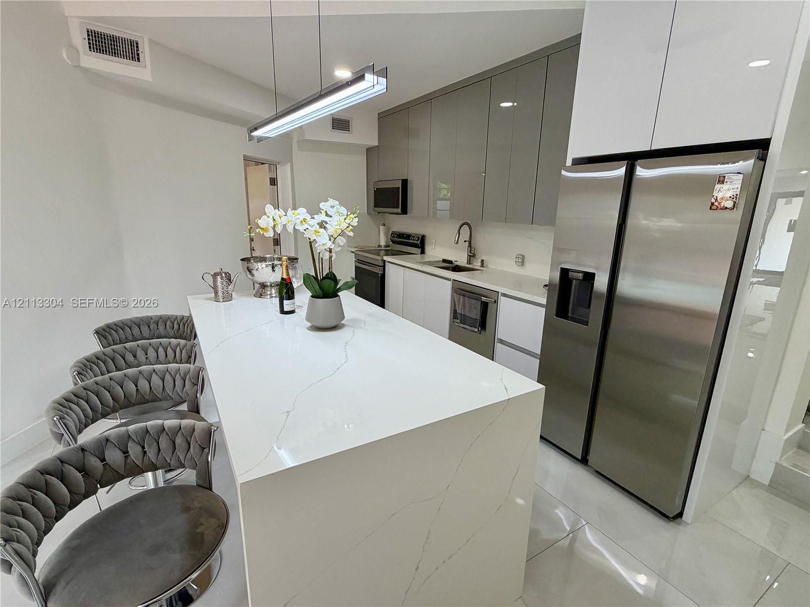 Kitchen featuring a waterfall-edge island with stone-finish countertop, high-gloss cabinetry, stainless steel appliances, and linear pendant lighting