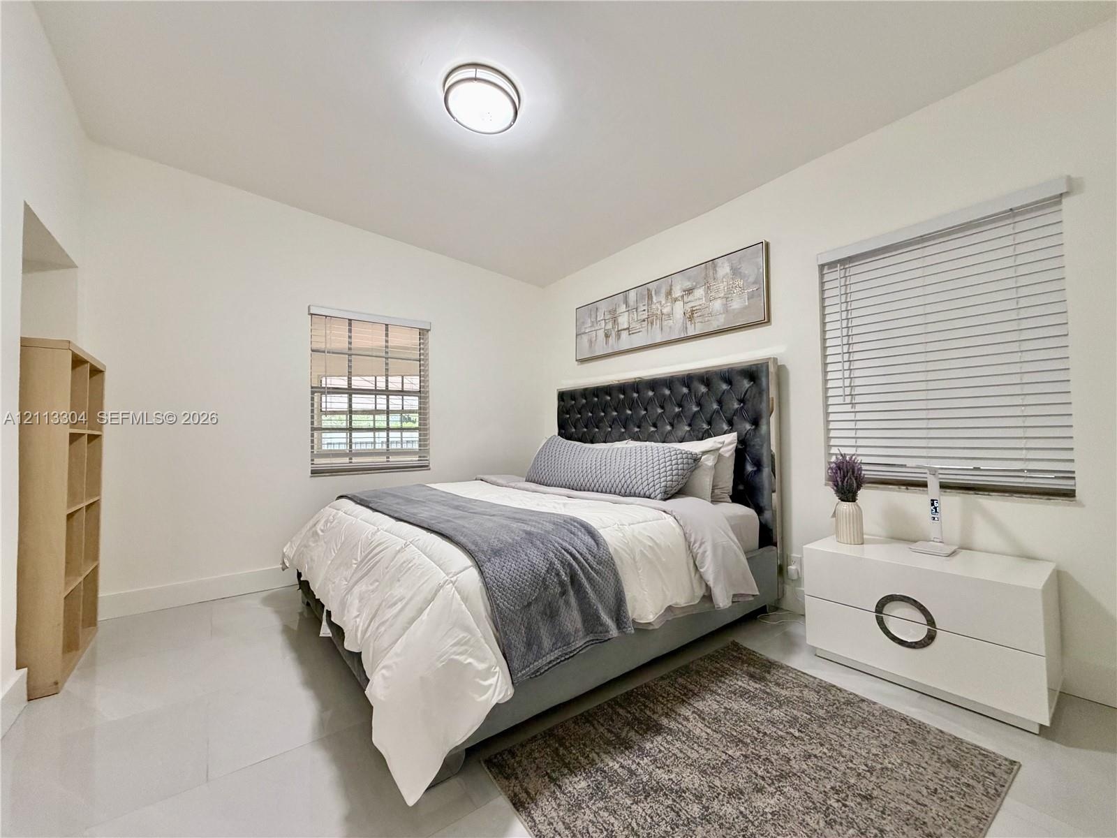 Neutral-toned interior featuring large-format tile flooring, recessed ceiling lighting, two windows with horizontal blinds, and a built-in open-shelvi
