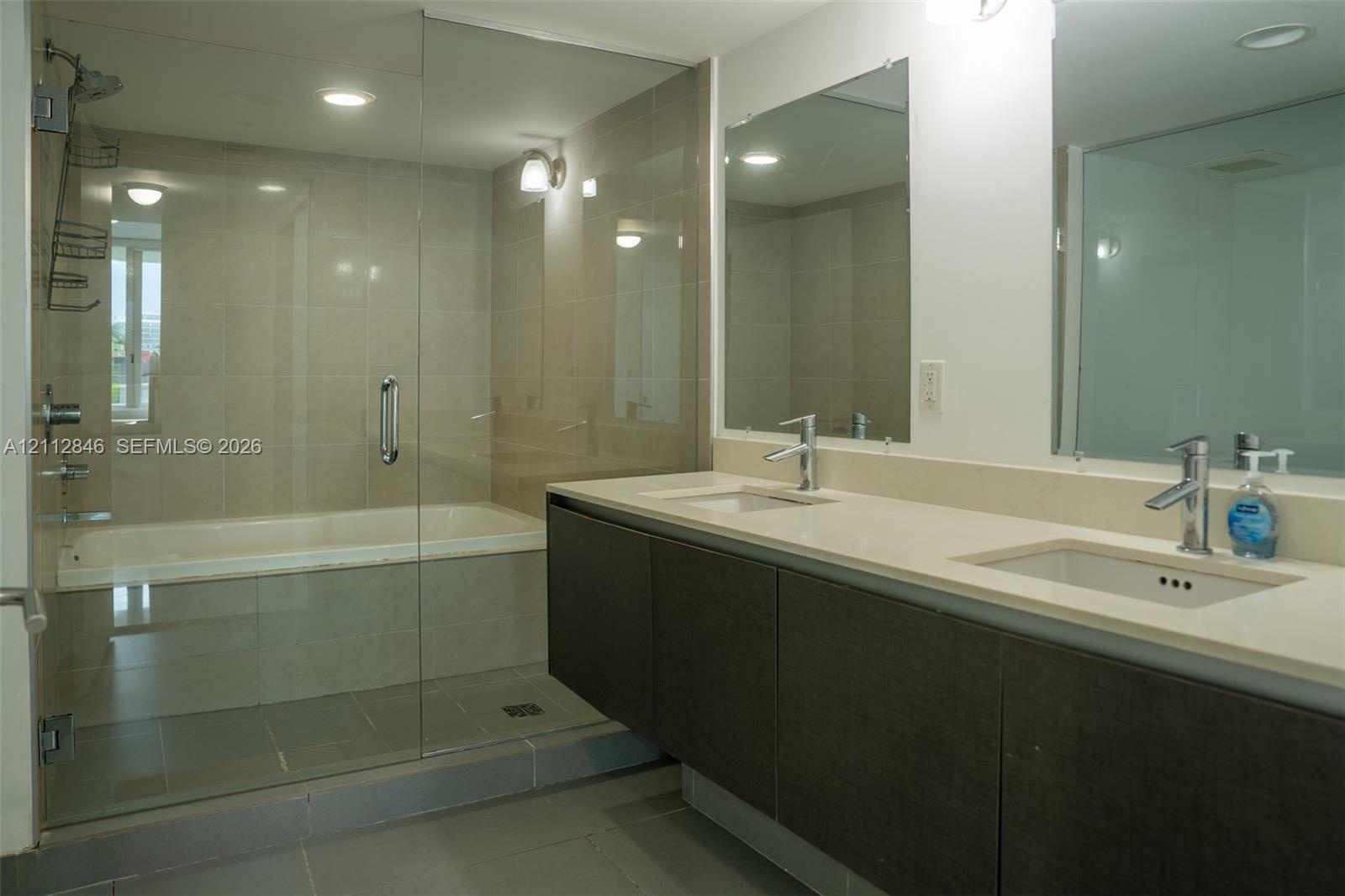 Dual-sink vanity with light-toned solid surface countertop and dark-finish cabinetry, complemented by a glass-enclosed wet room containing a soaking t