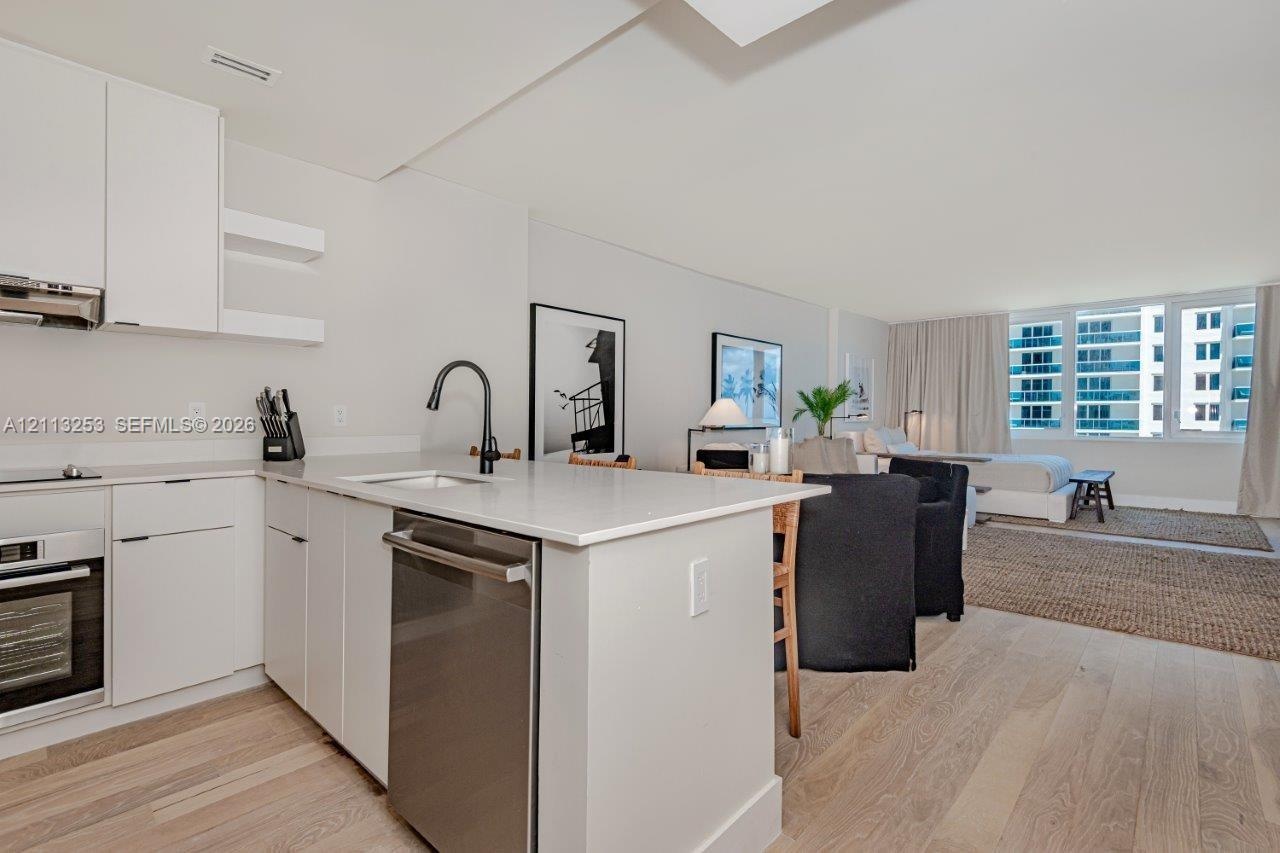 Kitchen featuring white flat-panel cabinetry, light-toned stone countertops, a stainless steel dishwasher, a matte black high-arch faucet, and light w