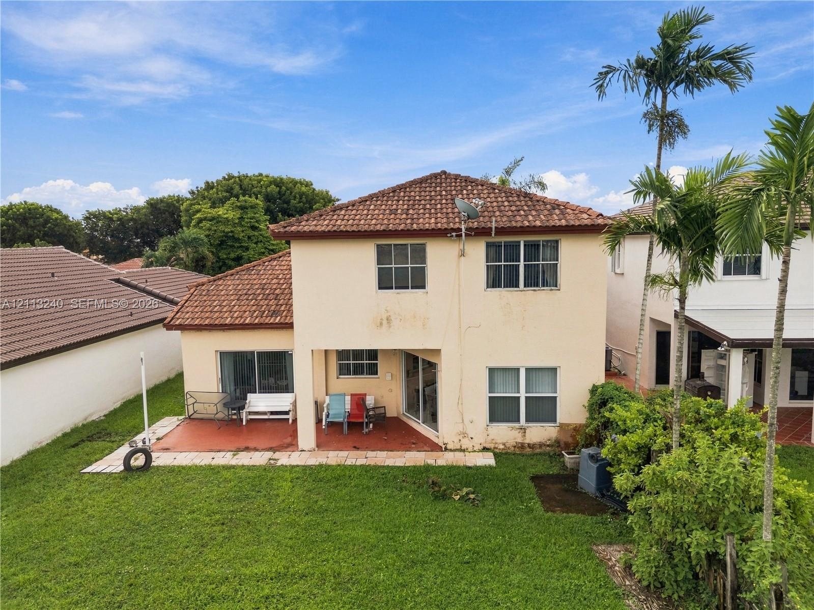Two-story exterior featuring a clay tile roof, stucco facade, covered rear patio, and expansive lawn with mature palm trees