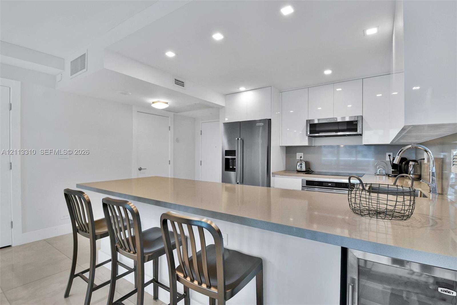 Kitchen featuring high-gloss white cabinetry, a neutral stone-surface peninsula, stainless steel appliances, recessed ceiling lighting, and tile floor