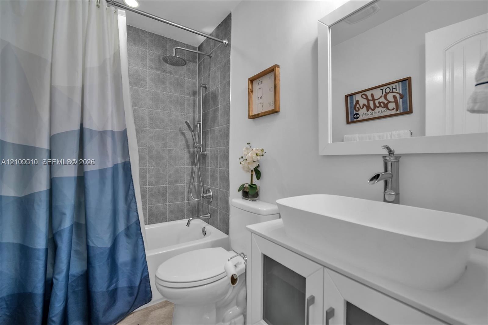 Bathroom featuring a white vessel sink with a chrome faucet, wall-mounted mirror with a white frame, shower with grey tile surround, adjustable handhe