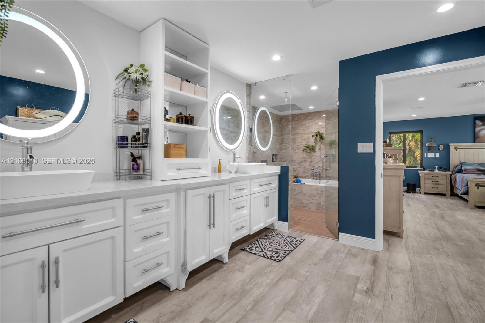 Dual white vanities featuring vessel sinks, integrated shelving, lighted circular mirrors, and a walk-in glass-enclosed shower with stone-tile surroun