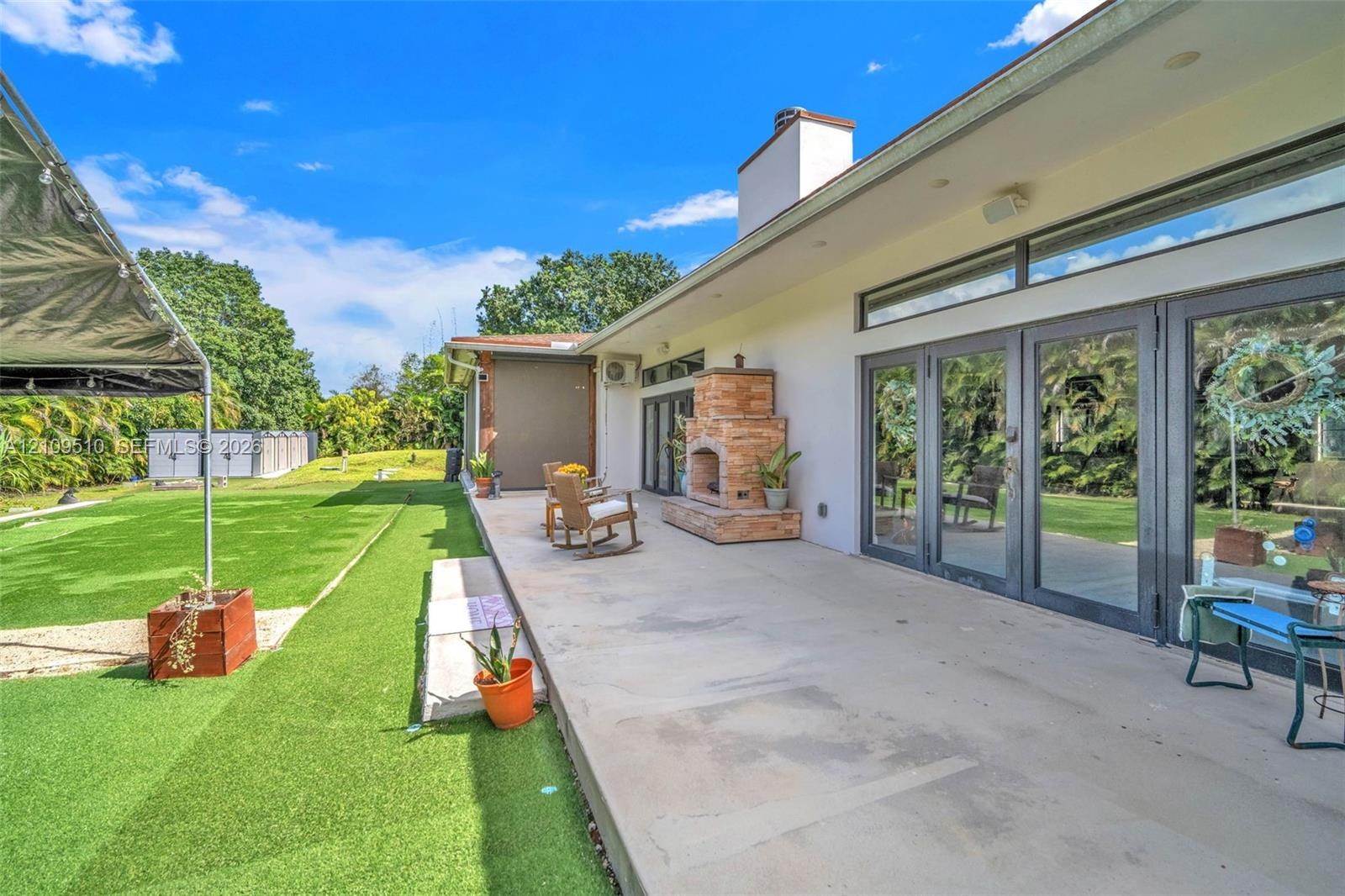 Spacious concrete patio featuring a built-in masonry outdoor fireplace, large glass multi-panel doors, and direct access to an expansive synthetic tur