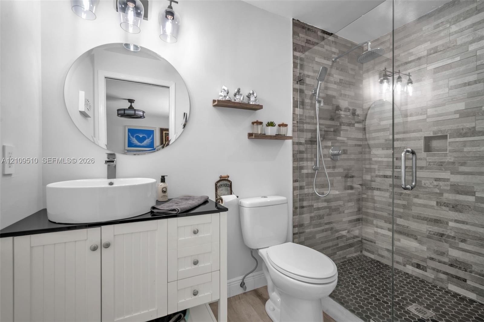 Bathroom featuring a white vanity with a dark countertop, a vessel sink, an oval wall-mounted mirror, and a walk-in shower with textured tile surround