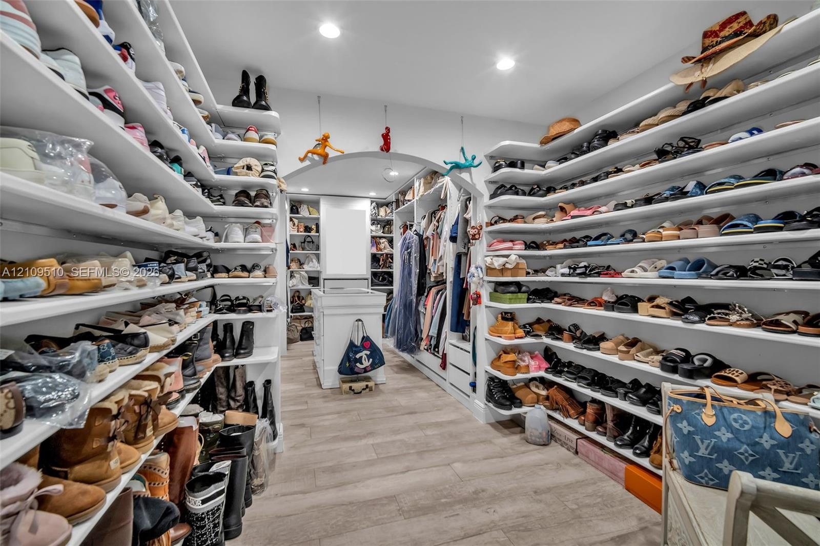 Custom walk-in closet featuring extensive built-in white shelving, recessed lighting, wood-finish flooring, and a central storage island