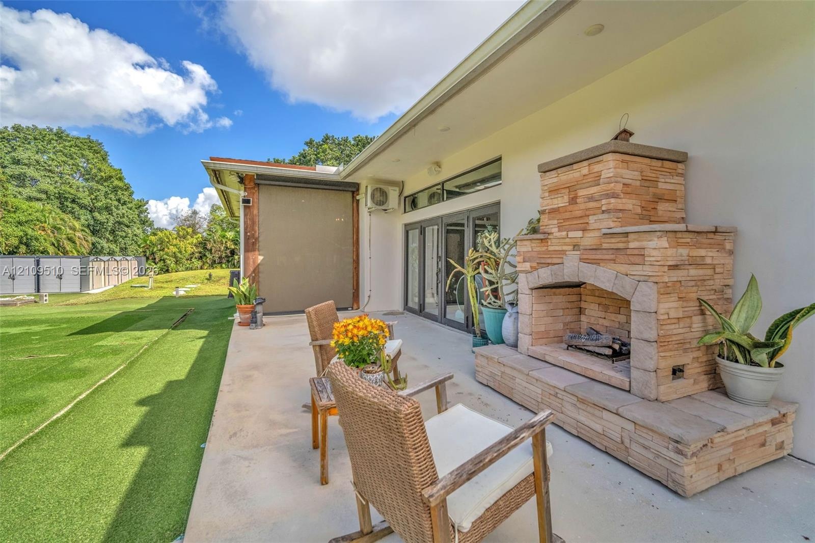 Covered concrete patio featuring a stacked stone fireplace with a raised hearth, glass multi-panel doors, and direct access to an expansive turf lawn