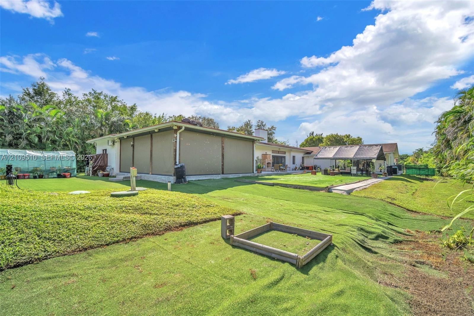 Expansive backyard featuring artificial turf, a permanent greenhouse structure, a covered patio with integrated privacy screens, and a timber-framed g