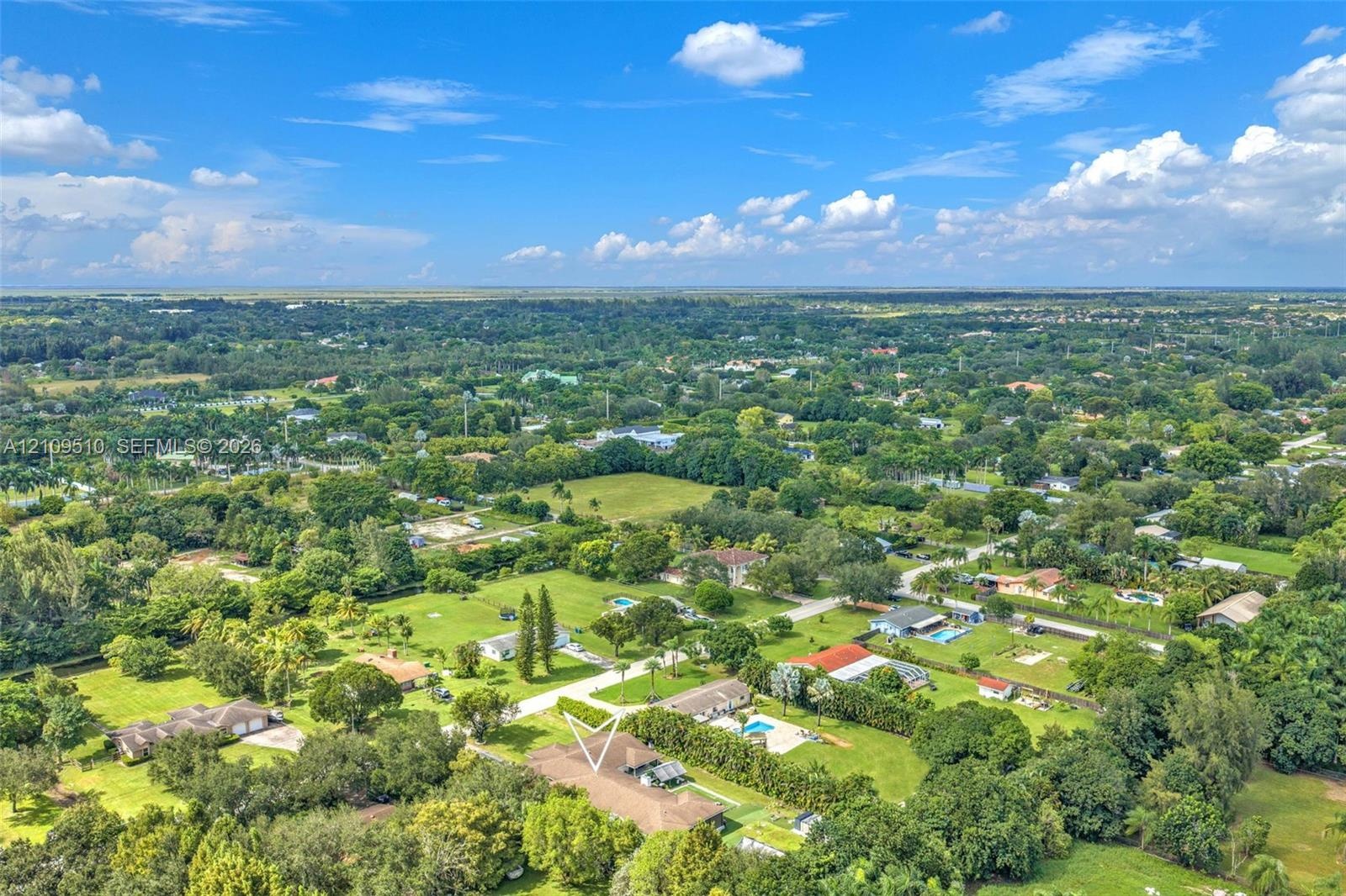 Aerial perspective of a sprawling property featuring a hip-roof residence, expansive grounds, mature landscaping, and significant perimeter vegetation