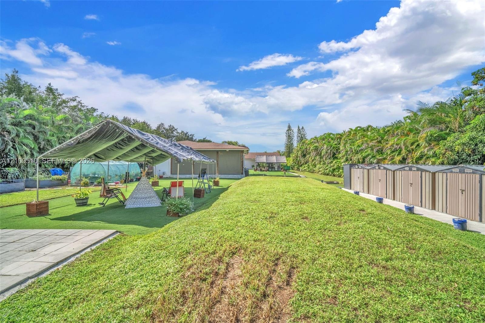 Expansive backyard featuring artificial turf, a metal-framed canopy, a semi-circular greenhouse, a concrete patio, and a row of external storage units