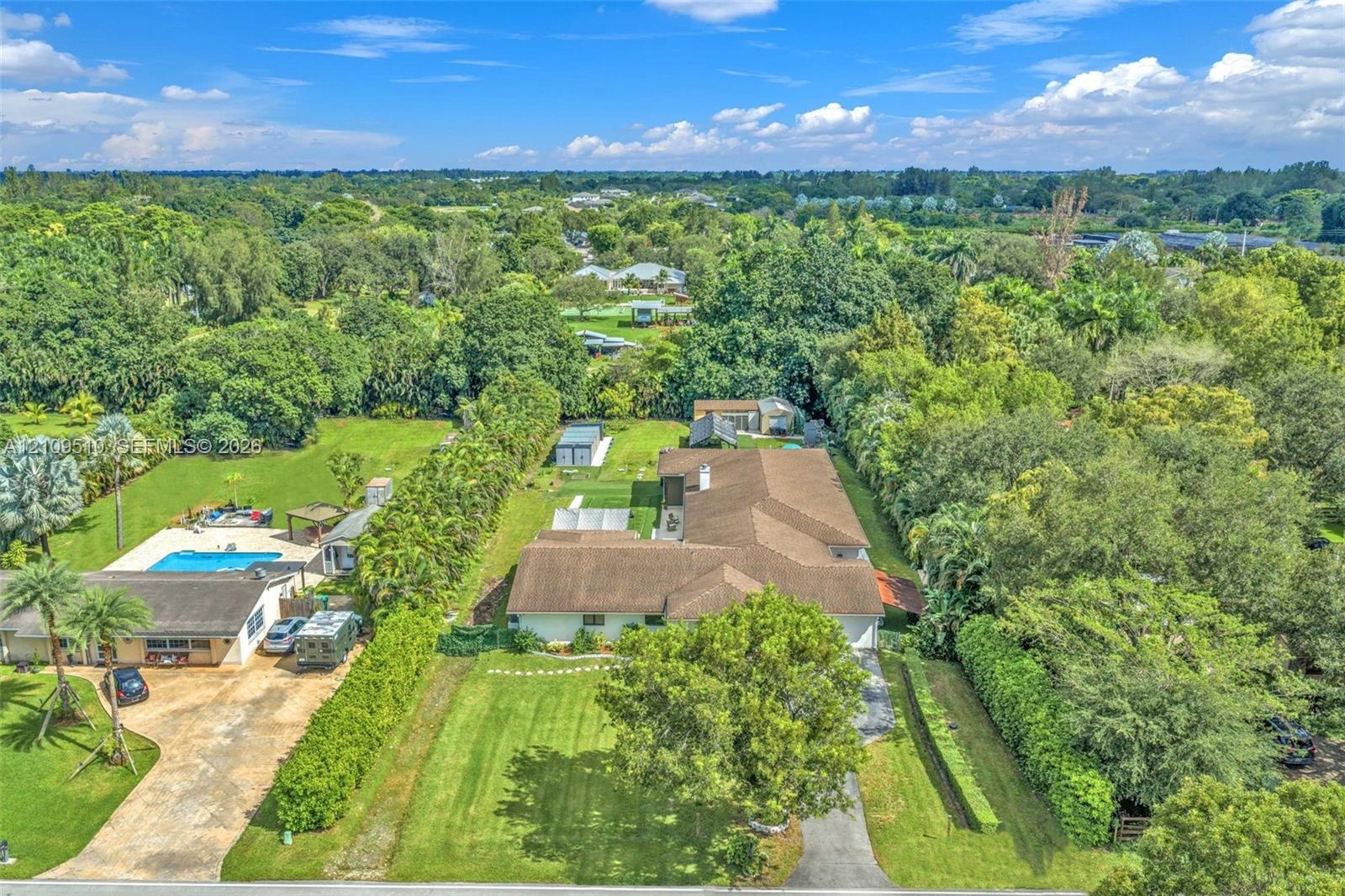 Aerial view of a sprawling property featuring an expansive lawn, mature tree canopy, multi-structured roofline, and an outdoor swimming pool with hard