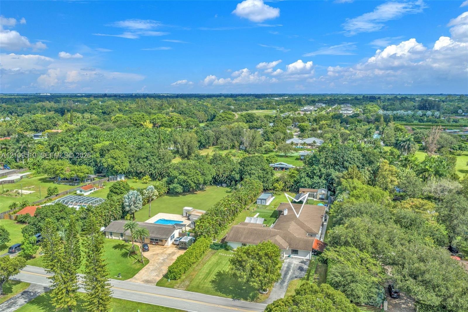 Aerial perspective of a detached residence featuring a multi-gabled roofline, concrete driveway, expansive lawn, and dense mature perimeter landscapin