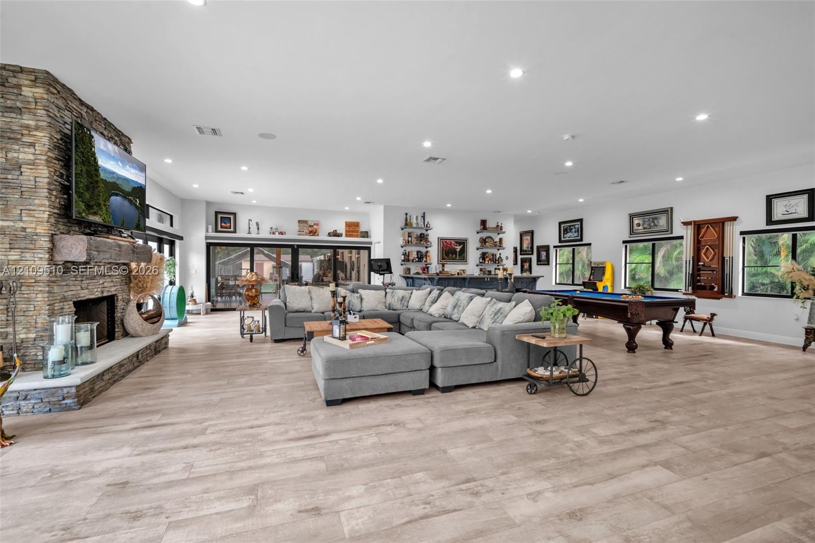 Open-concept living space featuring wood-finish flooring, floor-to-ceiling stone fireplace with a timber mantel, recessed lighting, and a built-in wet