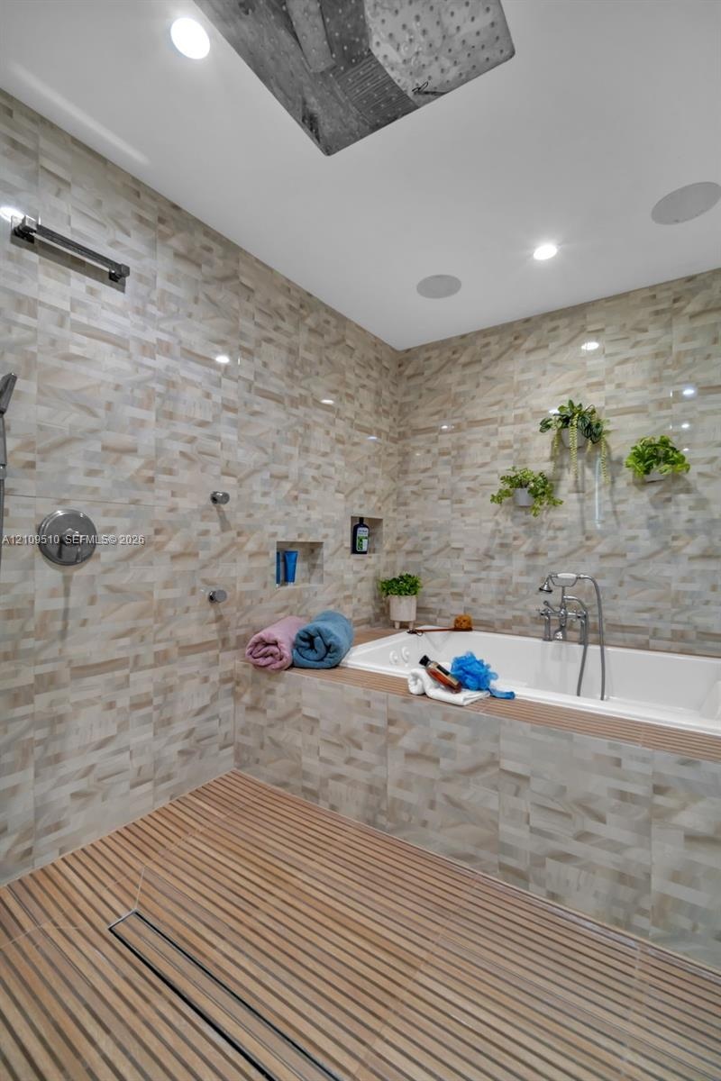 Wet room featuring a deep soaking tub, ceiling-mounted rainfall showerhead, linear floor drain, wood-slat flooring, and textured tile wall cladding
