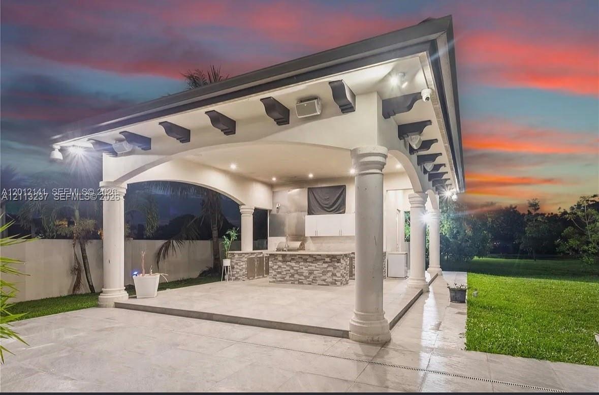Covered patio featuring architectural columns, exposed corbel brackets, recessed lighting, and an outdoor kitchen with stone-veneer cladding and tile 