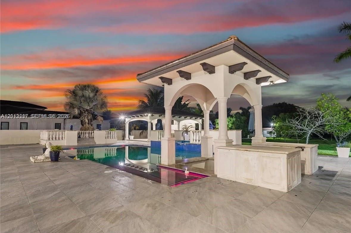Outdoor oasis featuring a swimming pool, covered pavilion with arched architectural detailing, stone-finish decking, and built-in masonry bar