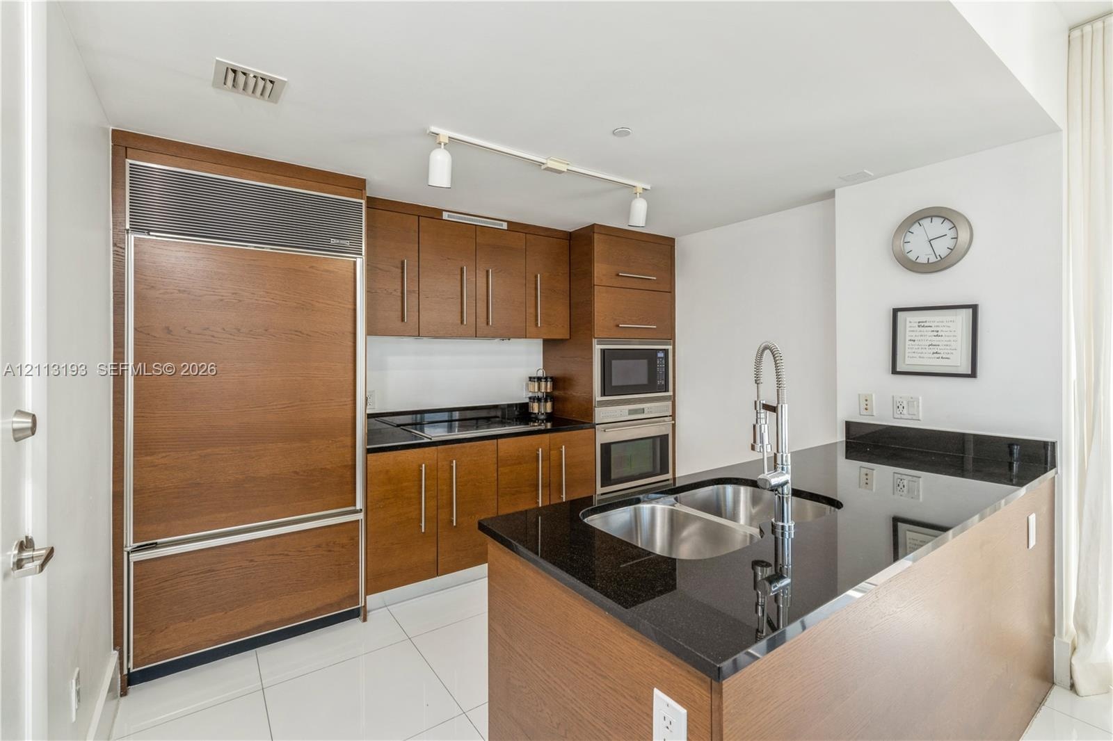 Kitchen featuring wood-finish cabinetry, black stone countertops, stainless steel appliances, dual-basin under-mount sink, and polished tile flooring